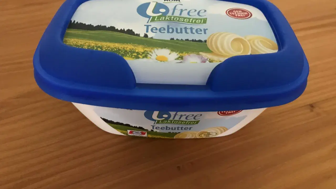 Eine Packung "Biofree Laktosefrei Teebutter" mit blauem Deckel steht auf einem Holztisch. Die Verpackung zeigt eine Sommerwiese mit Blumen und Butterflocken.