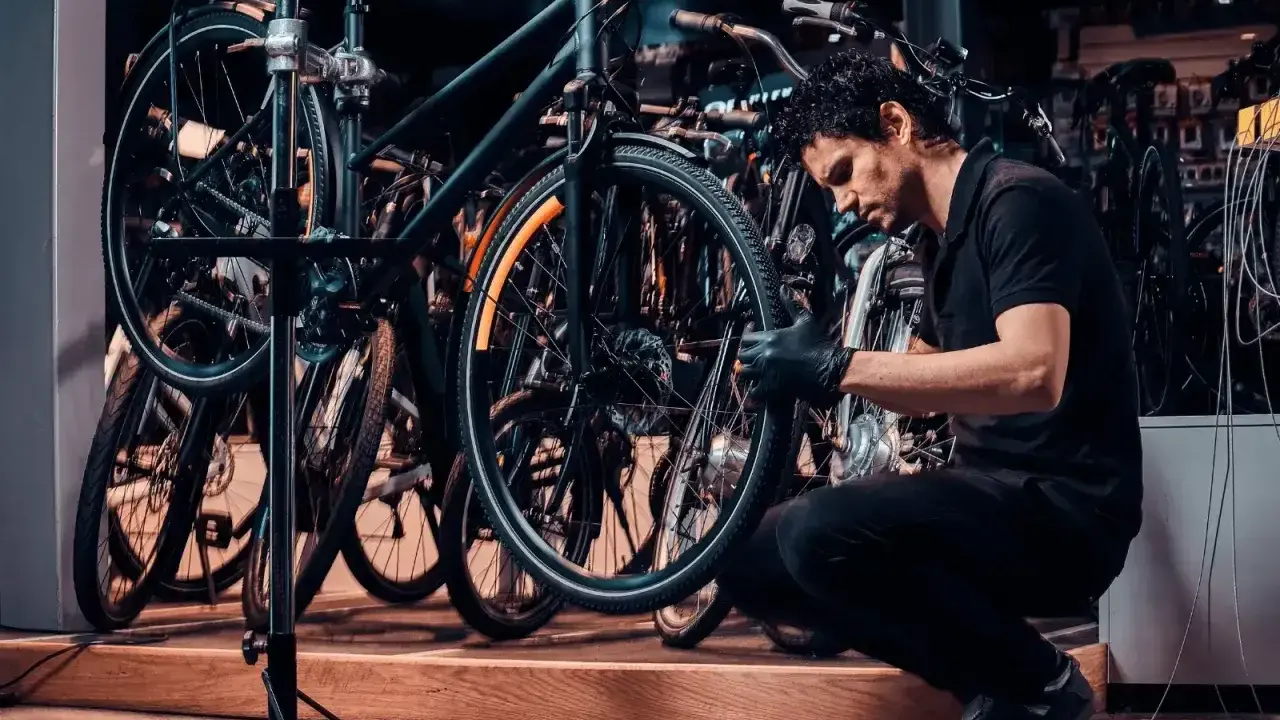 Mechanik w czarnej koszulce i rękawiczkach pokazuje, jak zdjąć tylne koło w rowerze, pracując w serwisie rowerowym.