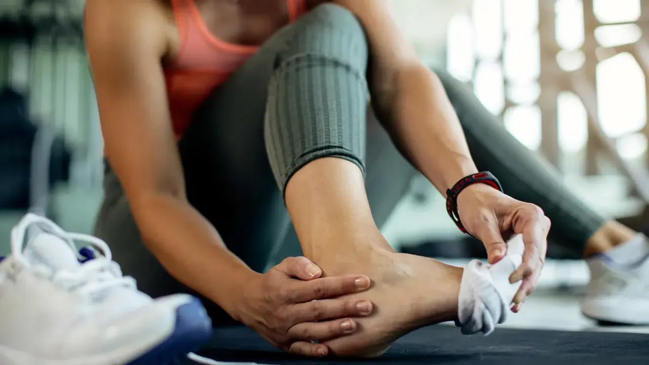 Kobieta w sportowym stroju masuje bolącą kostkę, obok leżą buty sportowe.