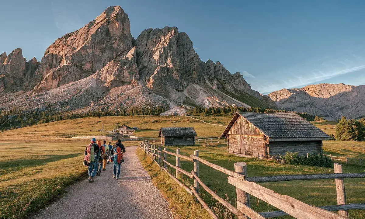 Piękna fotografia krajobrazowa Dolomitów. Grupa turystów wędruje ścieżką obok drewnianych chat, pod majestatycznymi szczytami górskimi.