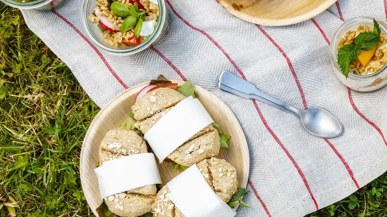 Idée pique-nique : petits pains garnis, salades en bocaux et muffins sur une nappe rayée, posée sur l'herbe.