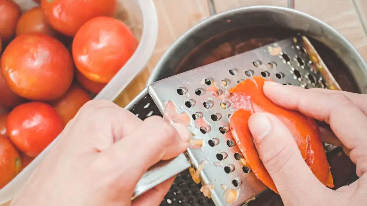 Ręce trą pomidora na tarce, przygotowując go do sosu. Obok stoi miska pełna świeżych pomidorów.