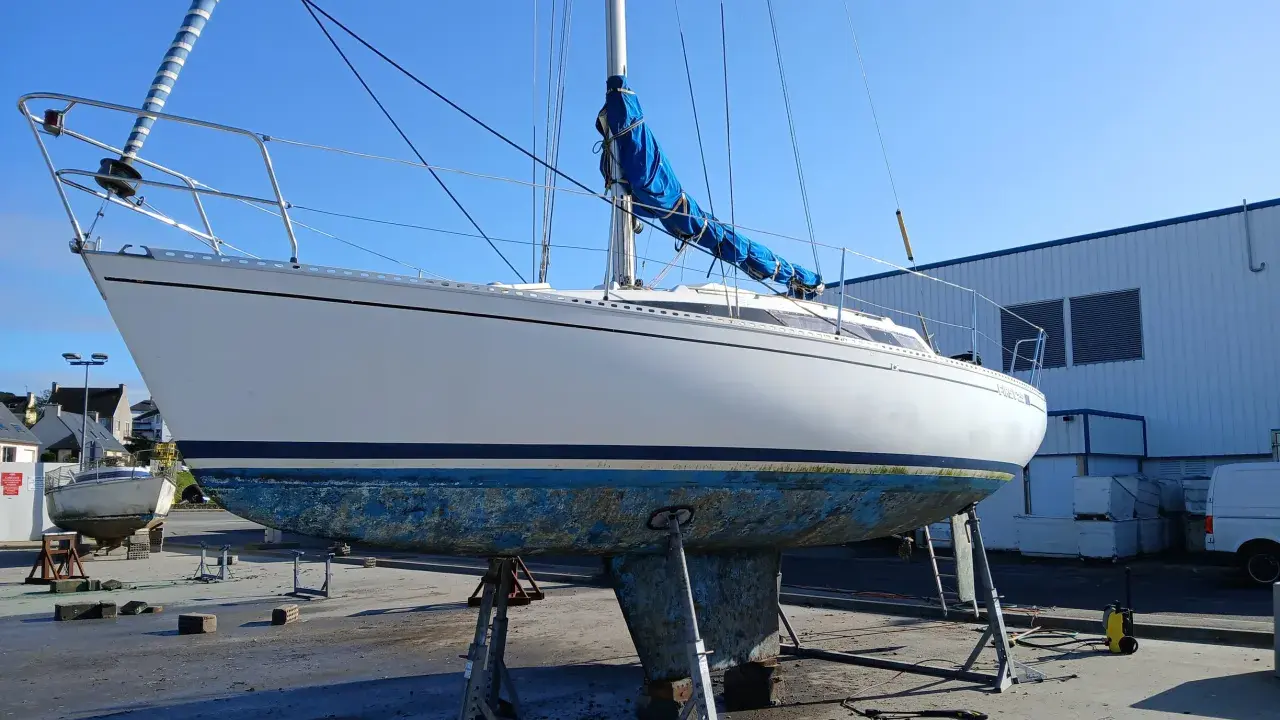 Voilier blanc sur bers, coque bleue usée, sous un ciel clair. Le mât est dressé, la grand-voile bleue enroulée. Premier 29 avis.
