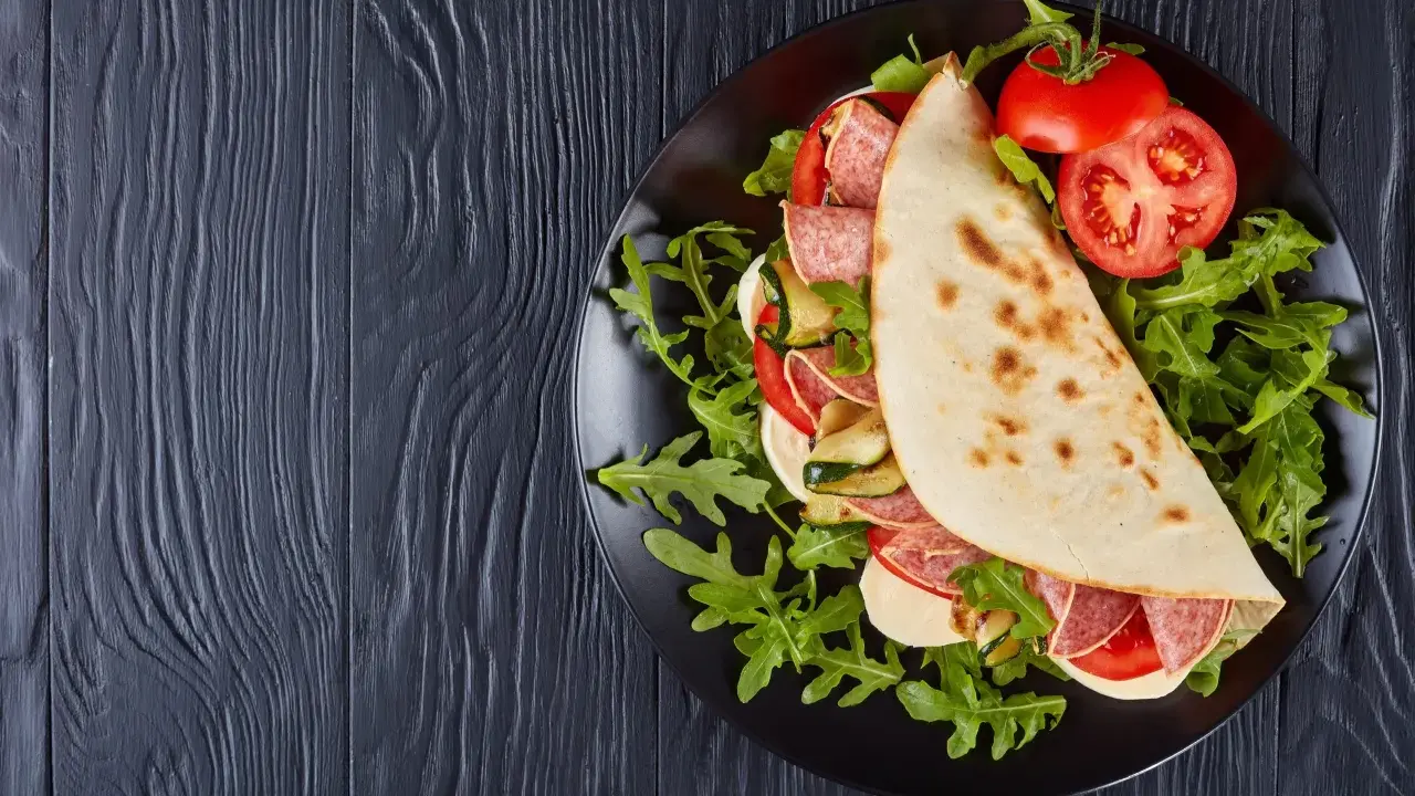 Piadina ripiena con salame, mozzarella, zucchine grigliate e pomodoro, servita su un piatto nero con rucola. Un assaggio della città di Rimini in un ristorante.