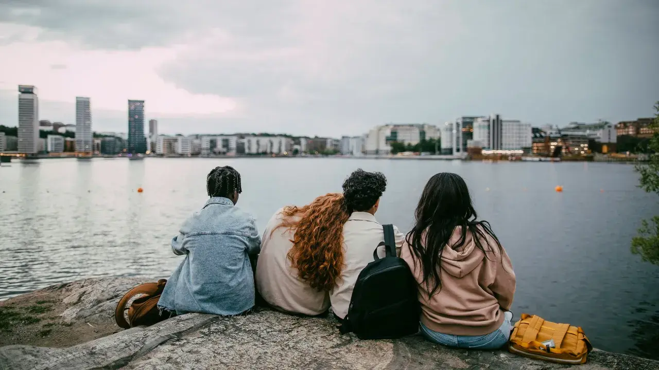 Three young people sit on rocks by the water, looking at a city skyline.