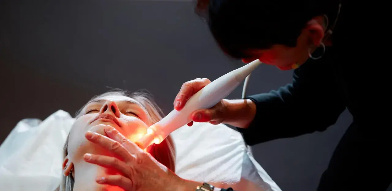 Une esthéticienne utilise un appareil Bloomea pour un soin du visage à la lumière rouge sur une cliente allongée.