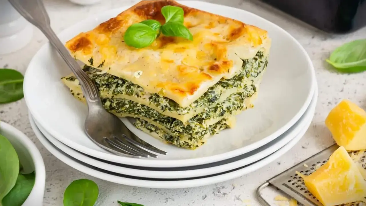 Ein Stück Spinatlasagne mit Käse überbacken, garniert mit Basilikum, auf einem Teller mit einer Gabel.