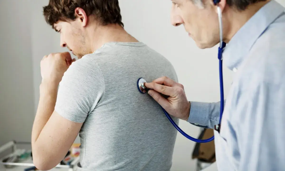 Arzt untersucht Patienten mit Stethoskop. Der Patient hustet, was auf Kurzatmigkeit hindeuten könnte.