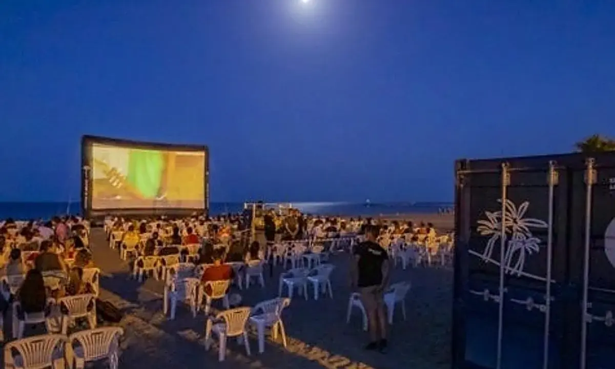 Noche de cine en el parque del cine Málaga. Público sentado en sillas en la playa, viendo una película en una pantalla gigante bajo la luna.