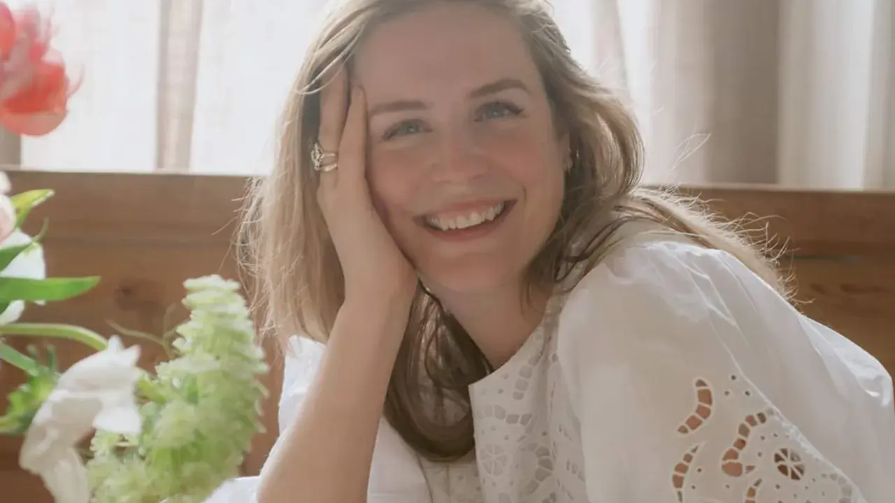 Glad kvinna med blont hår ler mot kameran, med handen mot kinden. En bukett blommor syns till vänster. Kanske ett foto från sofia wood blogg.