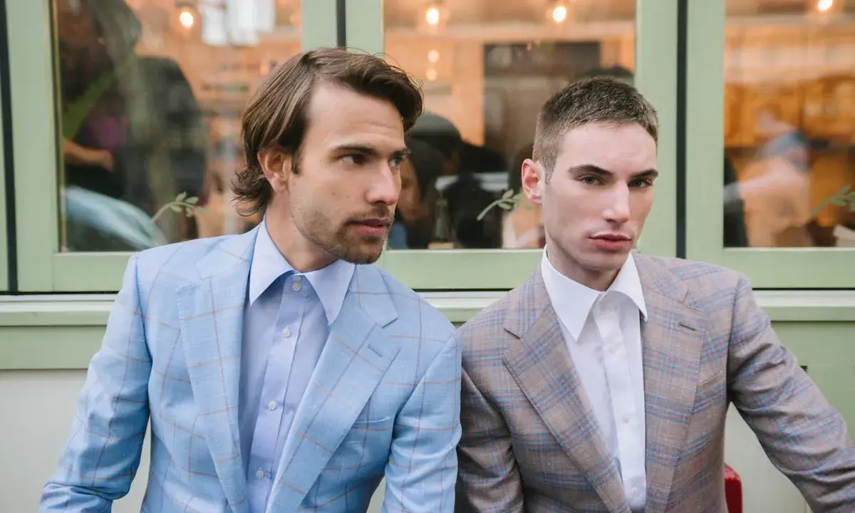 Two men in plaid suits with wide notch lapels sit side-by-side. The man on the left wears a light blue suit, the man on the right a tan suit.
