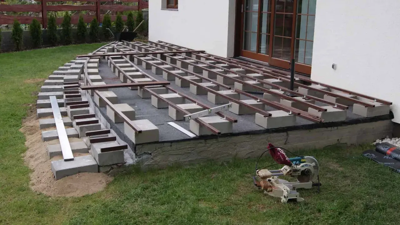 Budowa tarasu: betonowe bloczki i legary tworzą konstrukcję pod deski. Widać piasek, trawę i piłę.