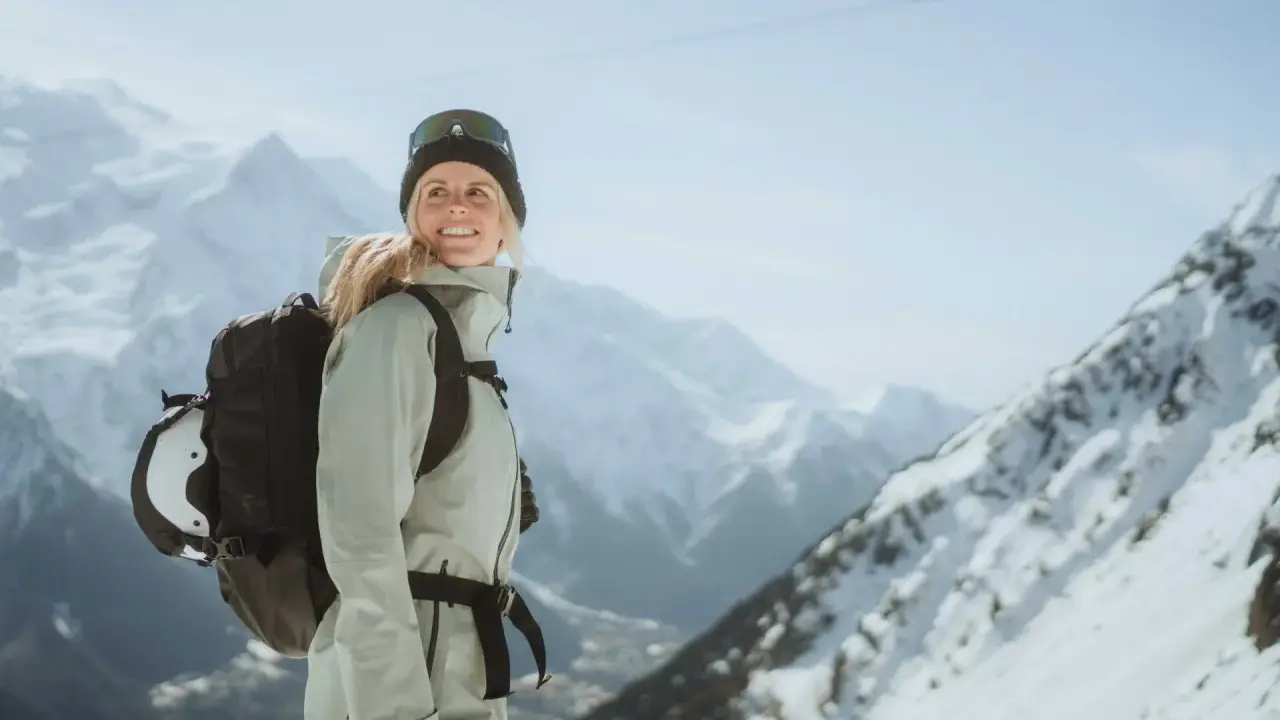 Kobieta w narciarskim stroju z plecakiem, do którego przymocowany jest kask, na tle gór.