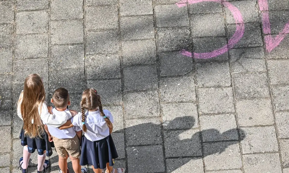Trójka dzieci w szkolnych strojach patrzy na napis "szkoła społeczna co to" narysowany kredą.
