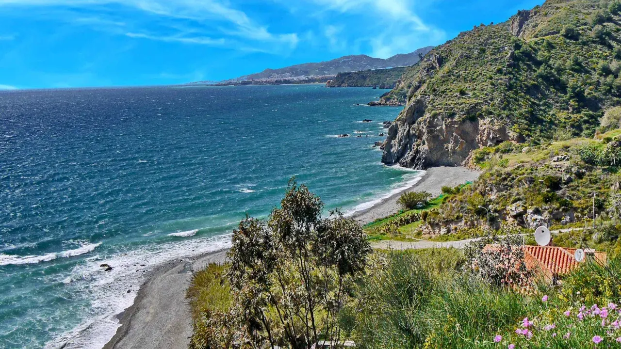 Ein malerischer granada strand mit felsigen Klippen, türkisfarbenem Meer und einer Stadt am Horizont.