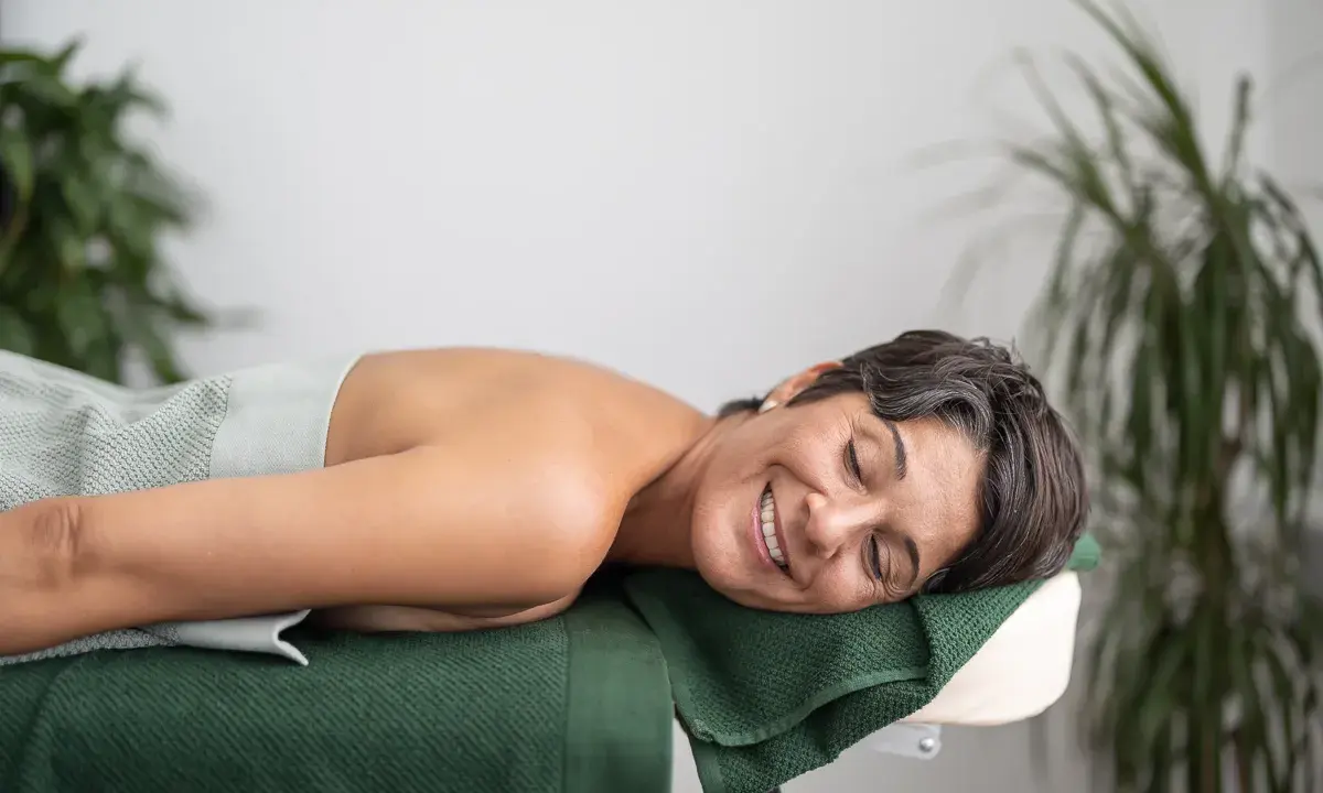En kvinna ler och njuter av att vila på en massagebänk, insvept i en ljusgrön handduk. Gröna växter i bakgrunden bidrar till en lugn atmosfär.