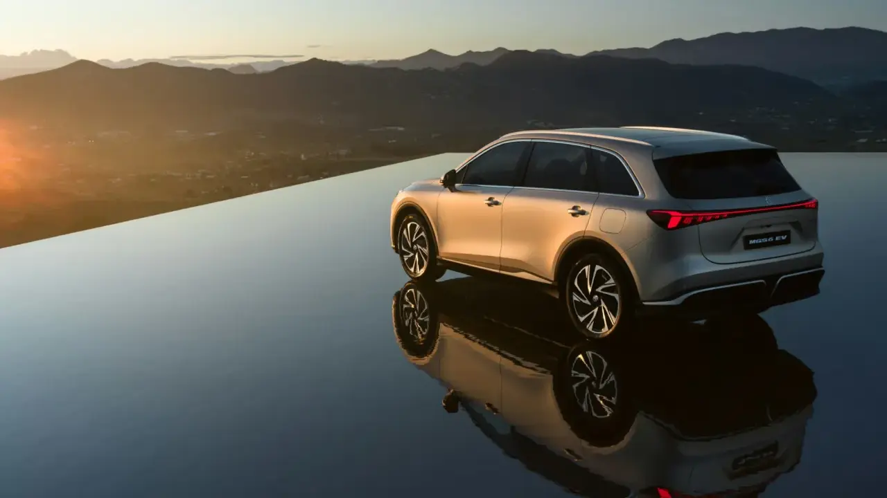 El MG6 EV plateado se refleja en una superficie de agua infinita al atardecer, con montañas al fondo.