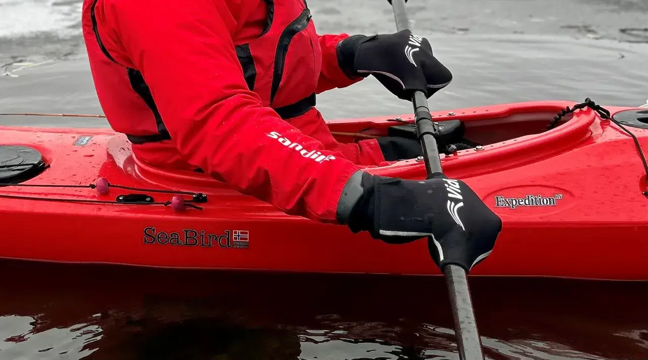 Person i röd jacka och svarta handskar paddlar kajak i kallt vatten.
