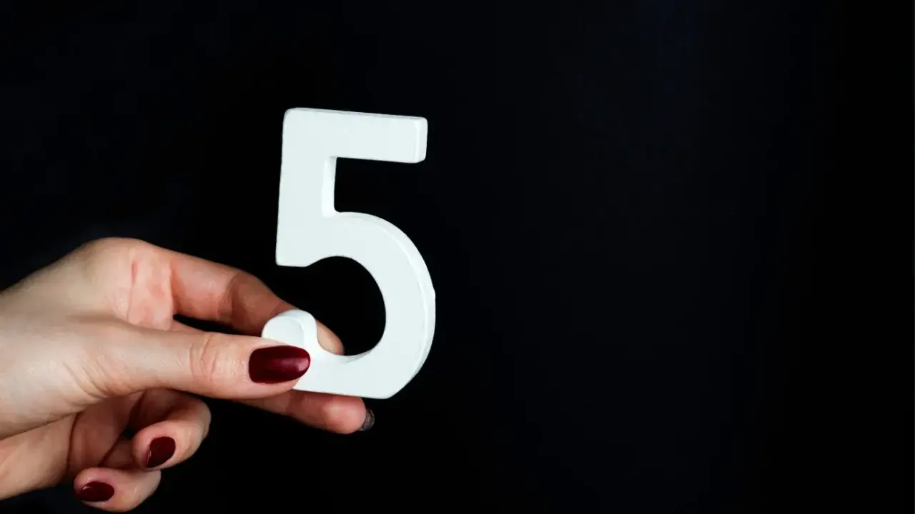 Dłoń z ciemnymi paznokciami trzyma białą cyfrę 5, symbolizującą numerologiczna 5 mężczyzna, na czarnym tle.
