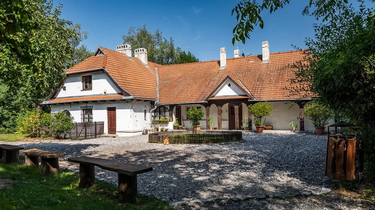 Urokliwy dom z czerwonym dachem w Bronowicach Małych, otoczony zielenią. Widać drewniane ławki i kamienny dziedziniec.