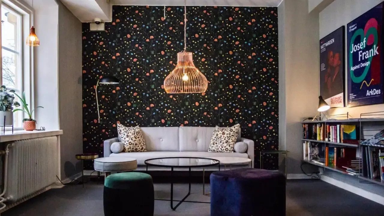 En mysig lounge med blommig tapet, en grå soffa, färgglada sittpuffar och en hylla full med böcker. En affisch för "Josef Frank - Against Design" hänger på väggen.