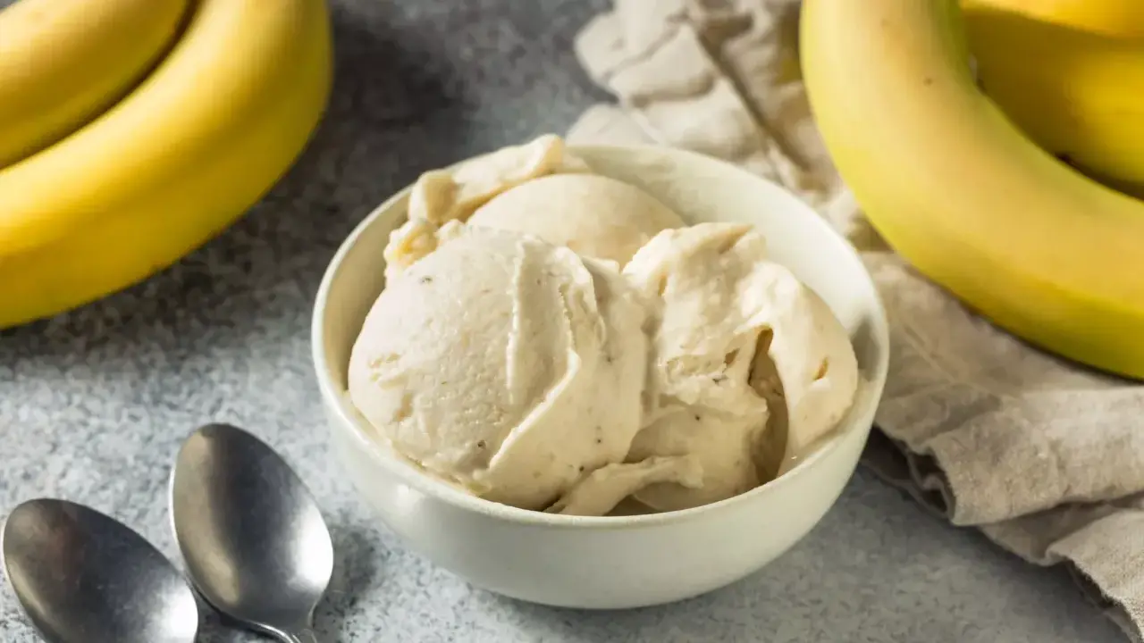 Un cremoso helado de platano servido en un cuenco blanco, con plátanos amarillos frescos al fondo.