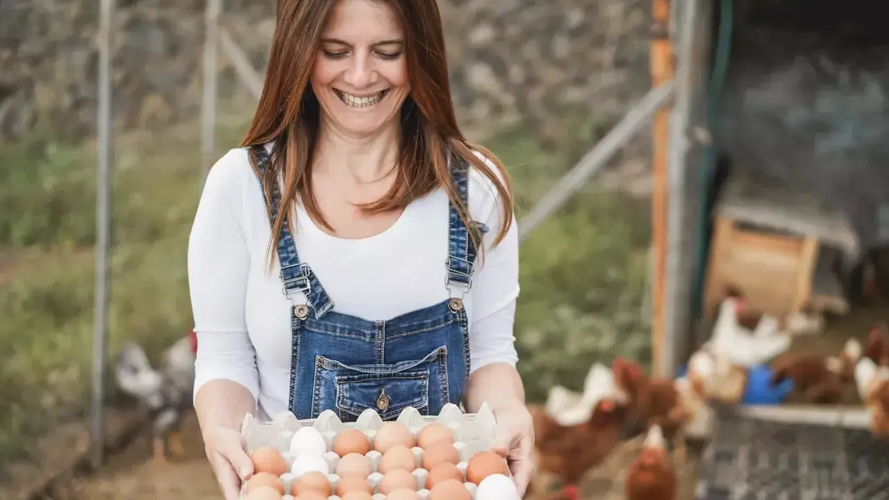Uśmiechnięta kobieta w ogrodniczkach trzyma tacę z jajkami. To świetne sprzedaż bezpośrednia przykłady świeżych produktów prosto od kury.