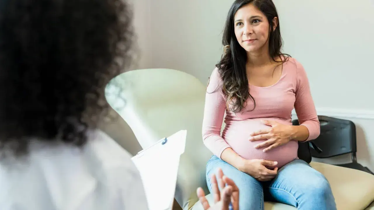 Schwangere Frau im Gespräch mit Arzt. Sie fragt sich, wie lange dauert eine einleitung, während sie ihren Bauch streichelt.