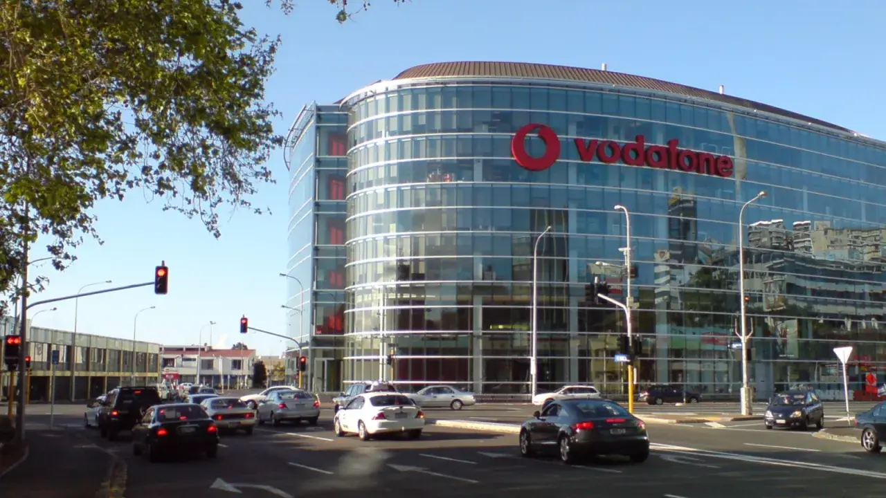 En modern glasbyggnad med stor Vodafone-logotyp, en livlig stadsgata med bilar och trafikljus. Vodafone Sverige syns tydligt.