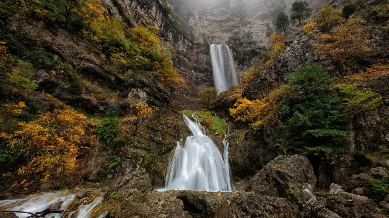 Cascada en un cañón rocoso, con árboles otoñales y niebla. El agua cae como un zafiro lagunazo, creando un espectáculo natural.