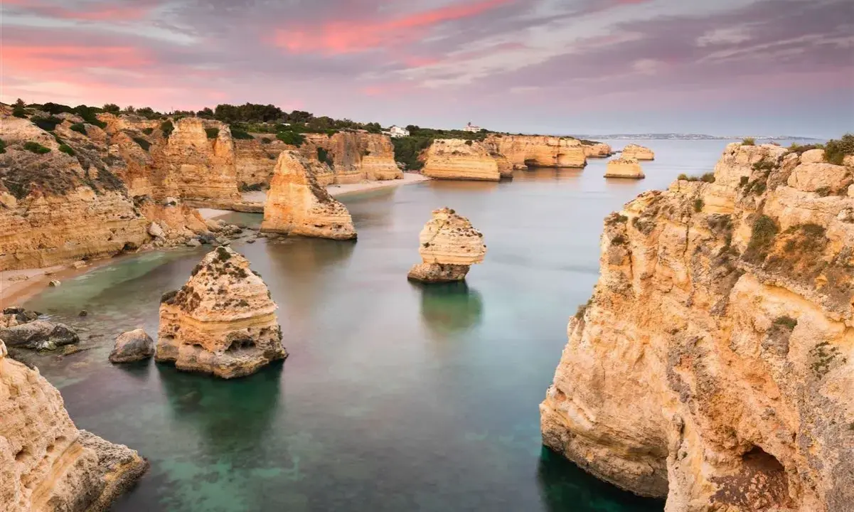 Felsformationen ragen aus dem ruhigen Wasser der **Lagoa, Portugal**, empor. Der Himmel ist in Rosa- und Orangetönen gefärbt, was auf einen wunderschönen Sonnenuntergang hindeutet.