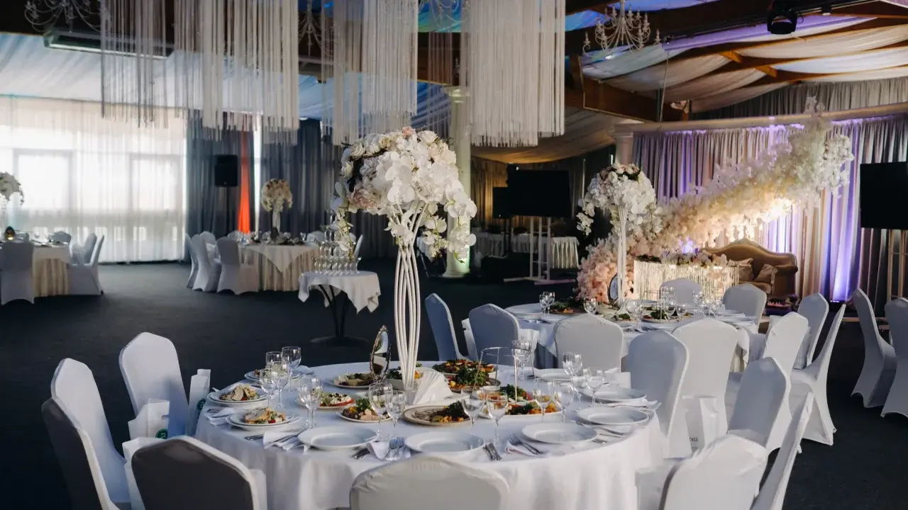 Elegancka sala weselna z białymi obrusami, kwiatami i oświetleniem tworząca magiczną atmosferę. To idealne miejsce na niezapomniane atrakcje na wesele.