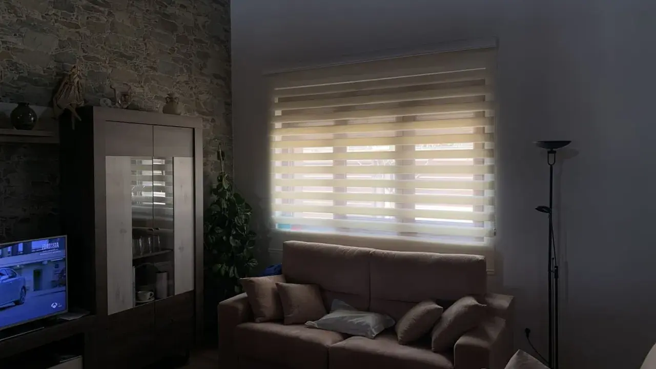 Salón acogedor con estores enrollables noche y día que filtran la luz, sofá, mueble y televisión.