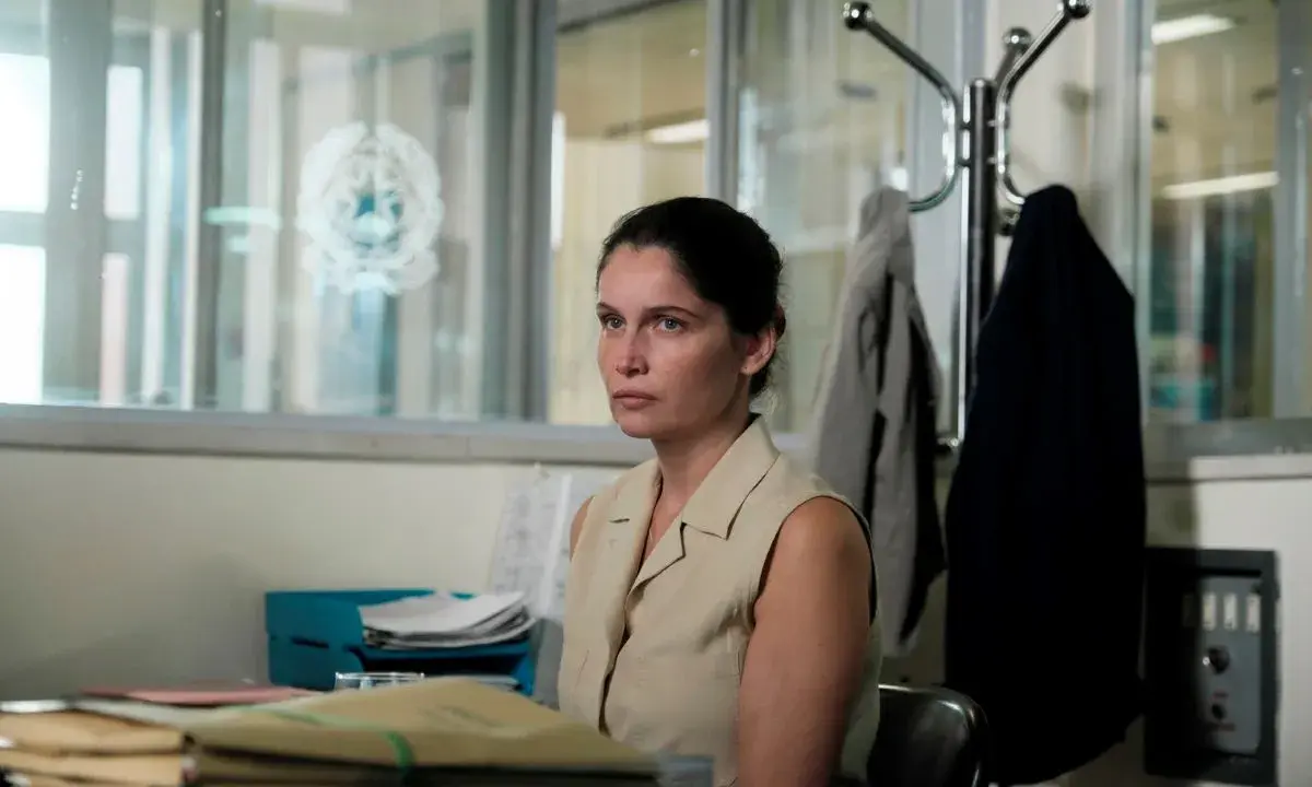Una donna con un'espressione pensierosa siede in un ufficio disordinato, circondata da fascicoli. Sembra l'inizio di una storia nera.