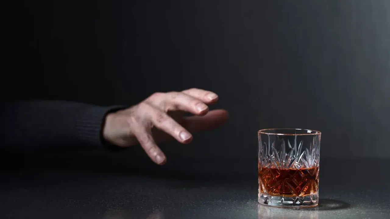 Mano tesa verso un bicchiere di whisky, un'immagine che evoca la domanda: quanto beve un alcolista al giorno?