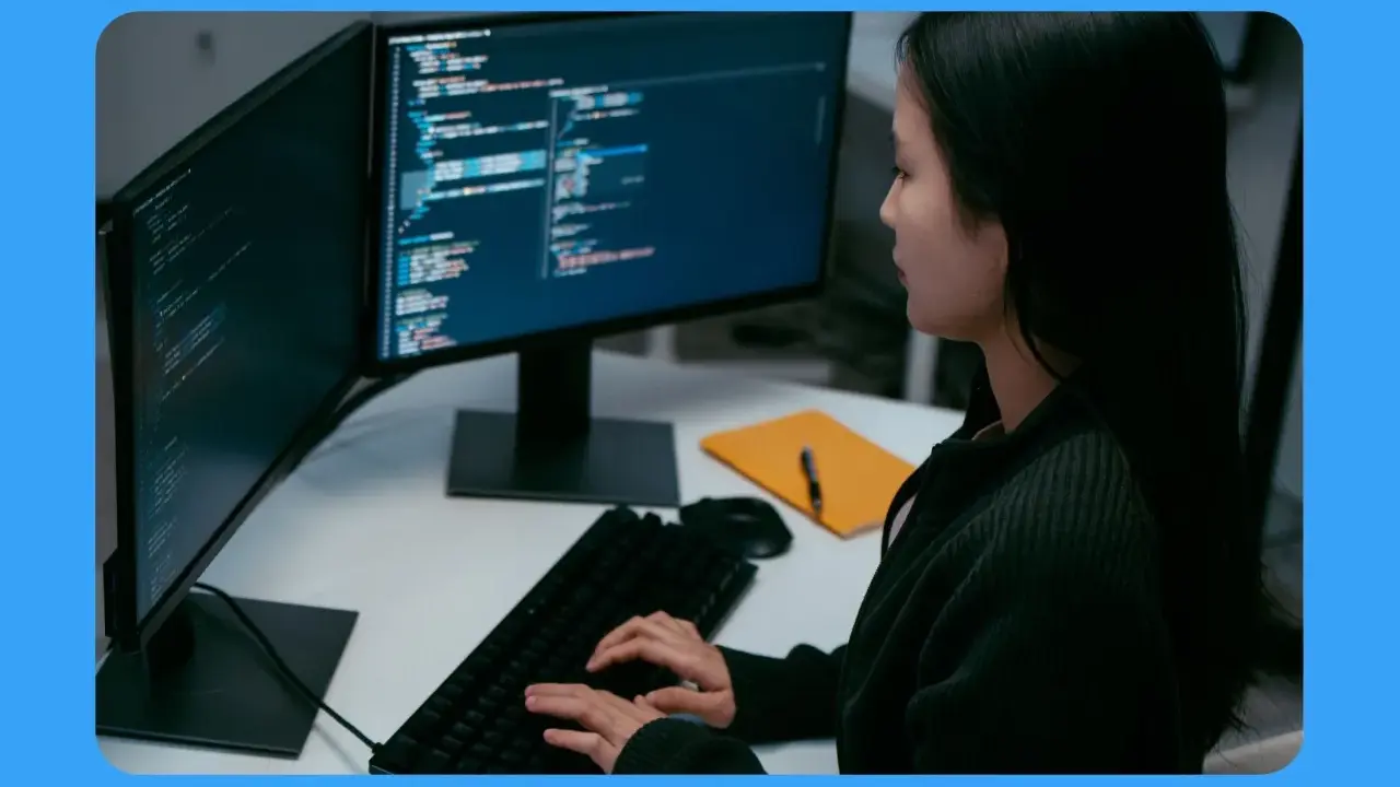 Jeune femme travaillant sur un ordinateur, avec deux écrans affichant du code. Sa position devant l'écran du bureau est concentrée.