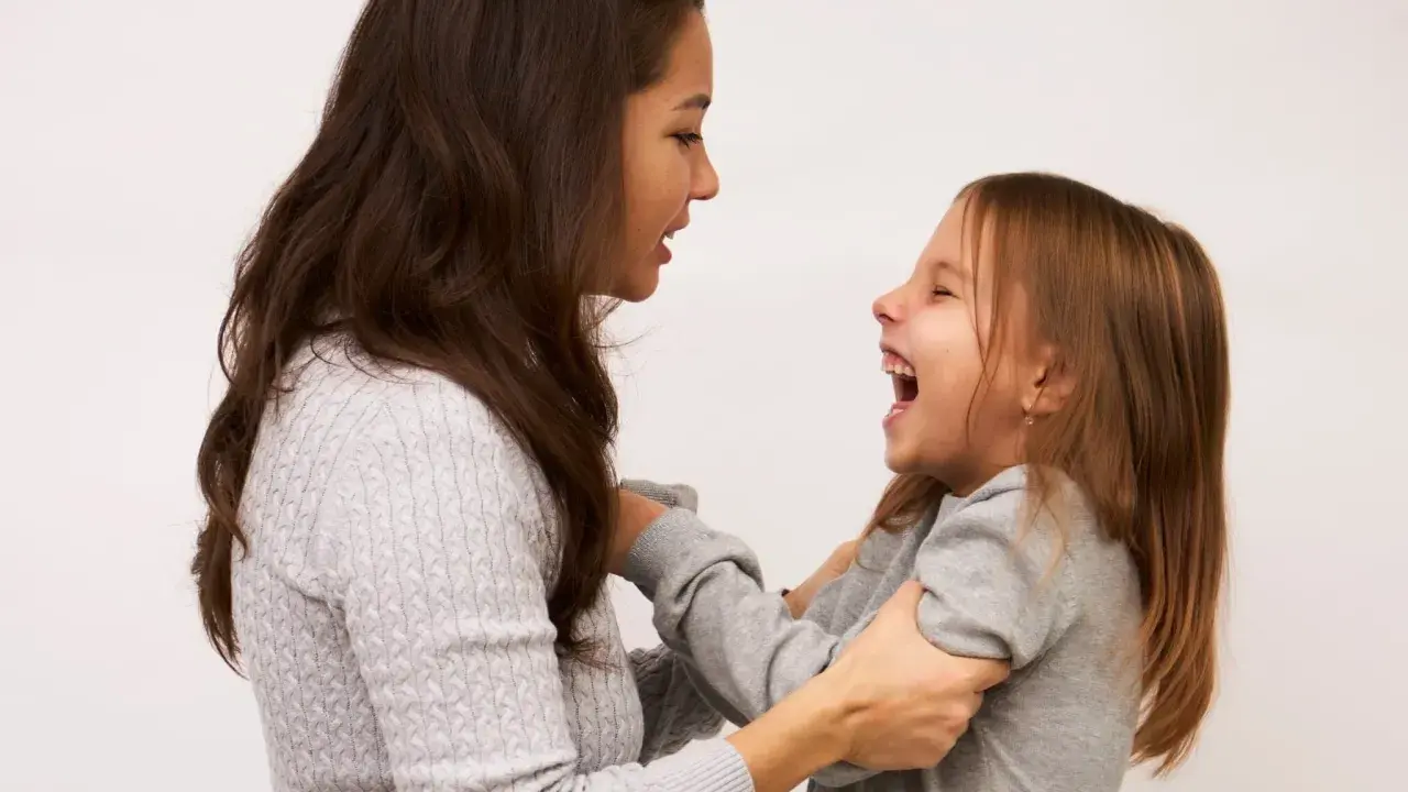 Mama śmieje się, łapiąc córkę za ręce. Dziecko radośnie szarpie się w nerwach, śmiejąc się głośno.