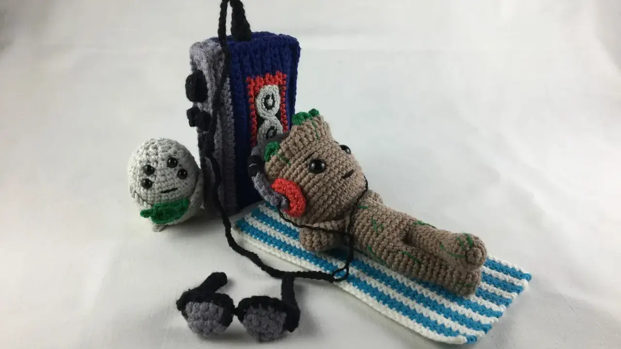 Amigurumi Baby Groot tumbado con auriculares junto a un reproductor de música y un pequeño personaje gris.