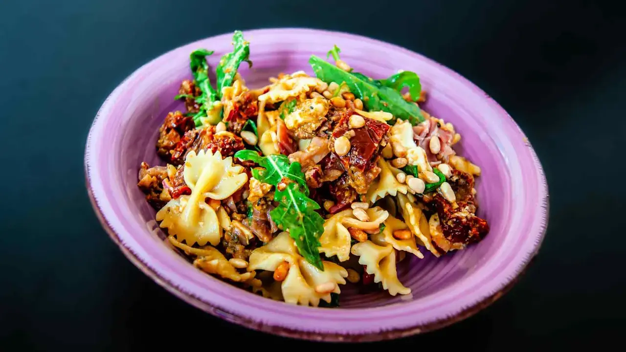 Ein köstlicher italienischer Nudelsalat mit Farfalle, sonnengetrockneten Tomaten, Pinienkernen und Rucola in einer lila Schüssel.