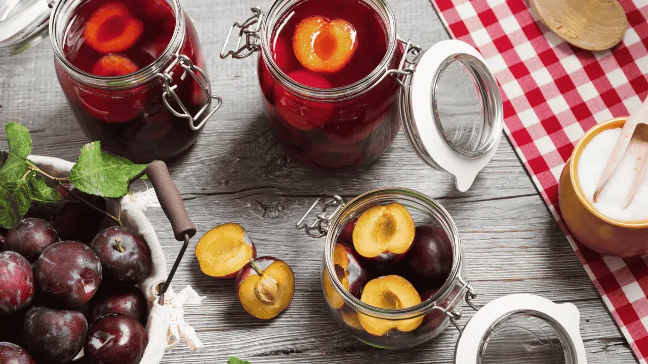 Frische Pflaumen werden für das Einkochen vorbereitet. Ganze und halbierte Früchte liegen neben Gläsern mit eingelegten Pflaumen.