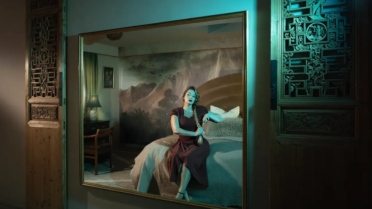 Elisa Miller sits on a bed, her reflection captured in a large mirror. The room features ornate wooden panels and a dramatic mountain mural.