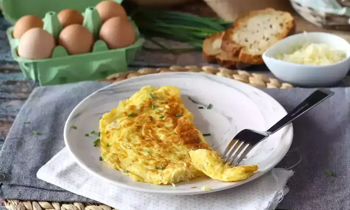 En gyllene omelett med ost, toppad med gräslök. Bredvid syns ägg, rostat bröd och mer ost. En perfekt start på dagen!