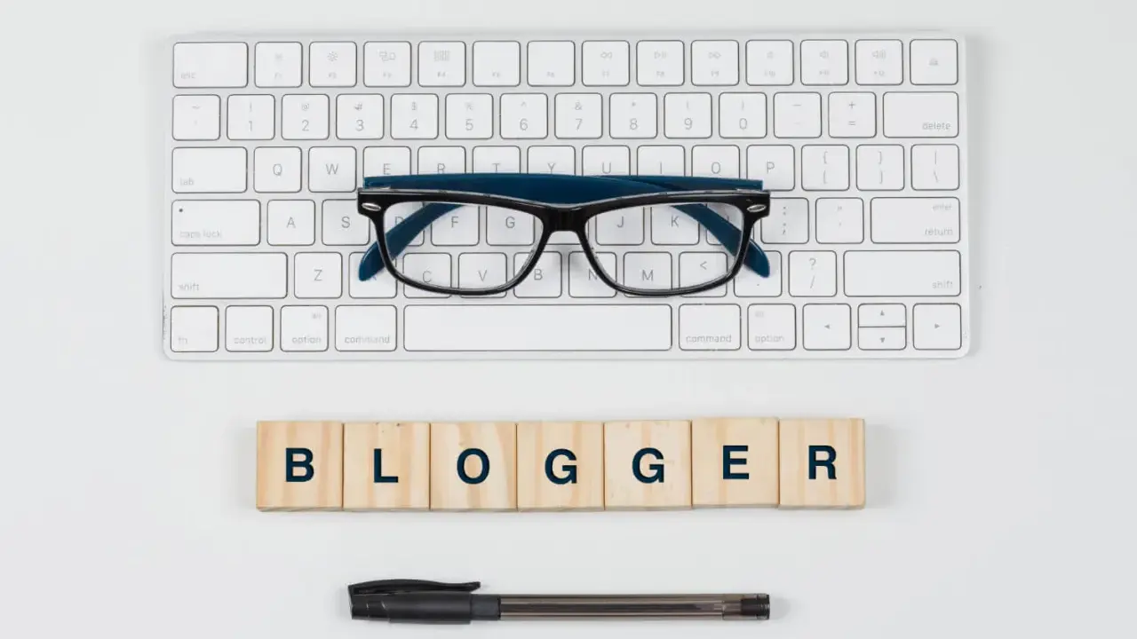 Okulary, klawiatura i drewniane klocki z napisem "BLOGGER" - idealne narzędzia do pisania bloga po angielsku.