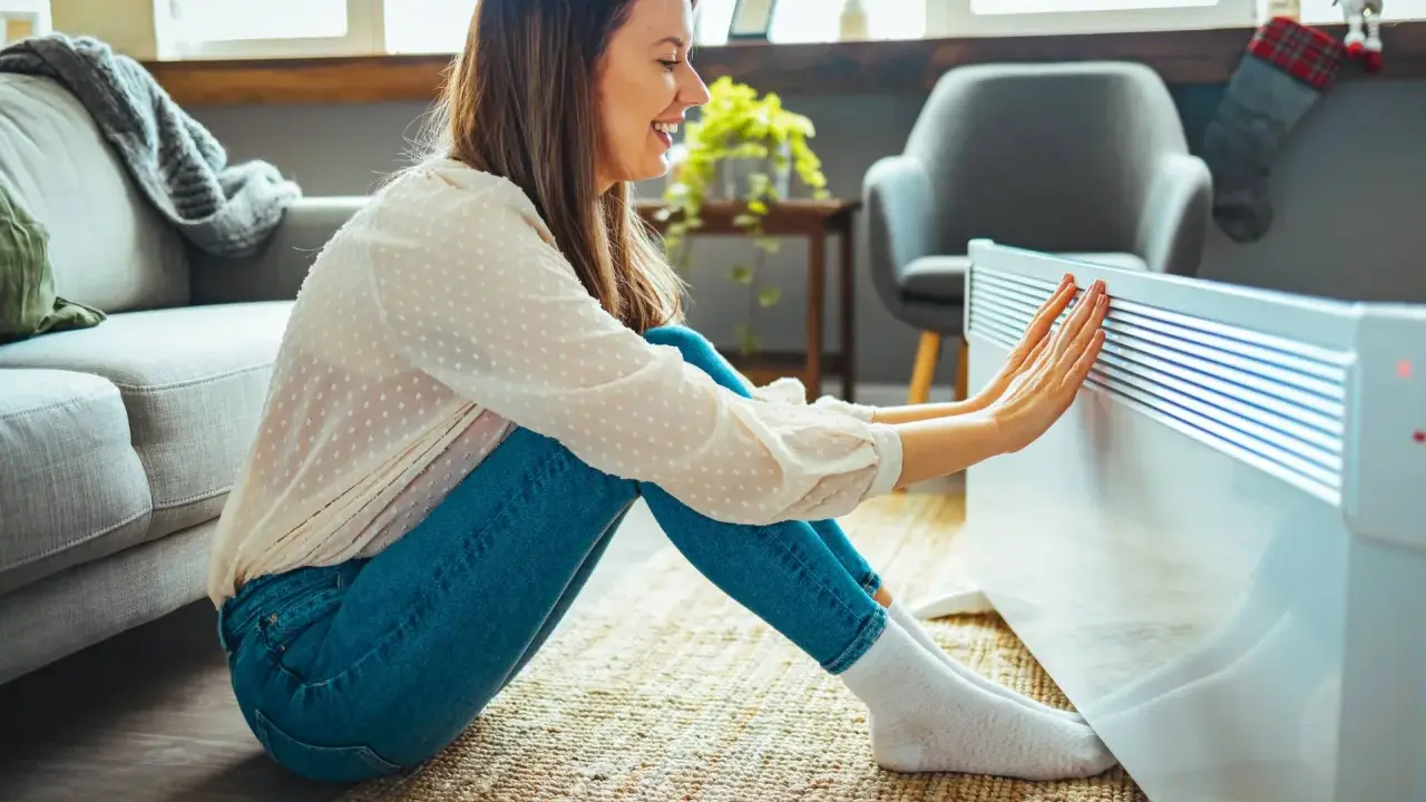 Mujer sonríe mientras calienta sus manos en un radiador, disfrutando de la temperatura ideal en casa en invierno.