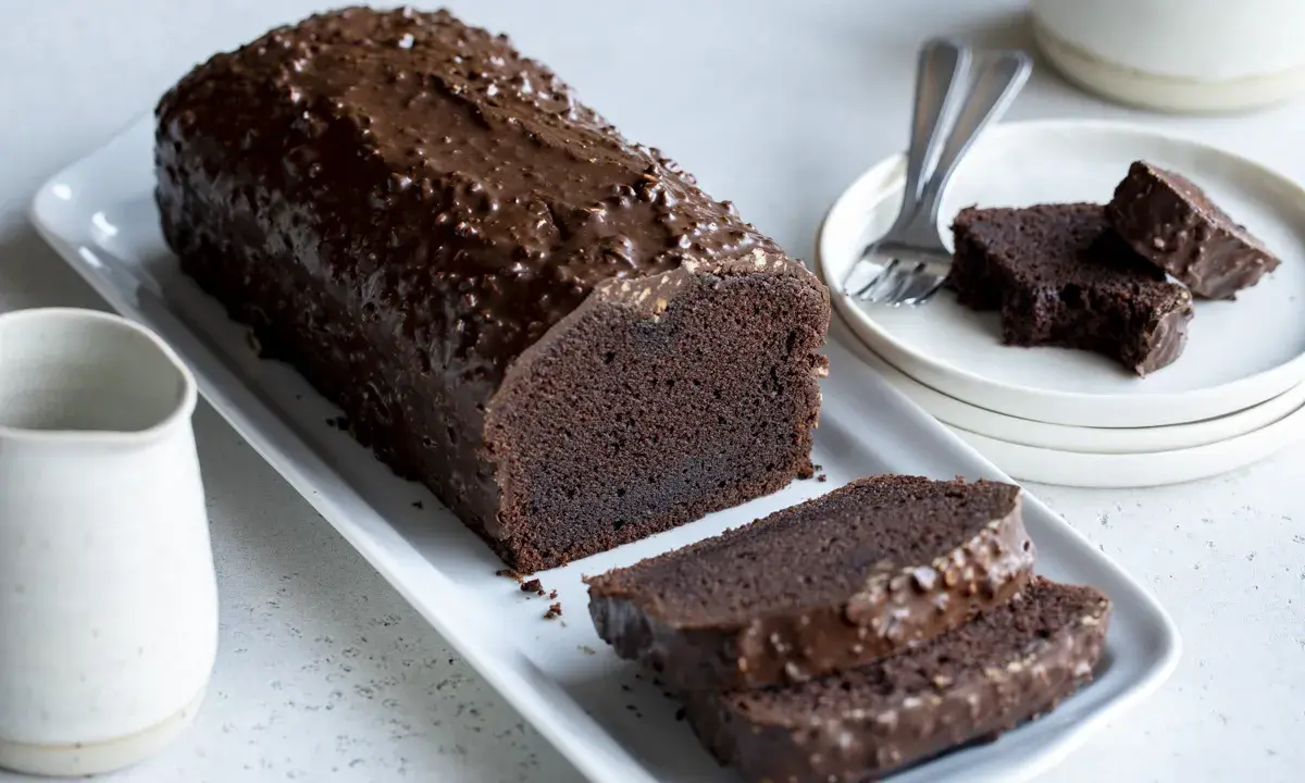 Un généreux gâteau au chocolat haut, nappé d'un glaçage croquant et parsemé de noisettes. Deux tranches sont déjà coupées, prêtes à être dégustées.
