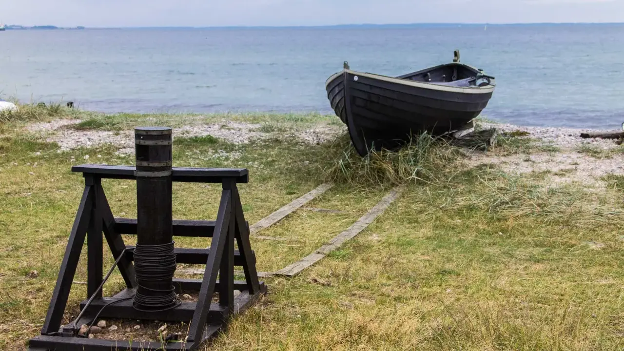 A dark wooden boat rests on a grassy shore next to a winch and wooden tracks leading to the sea.