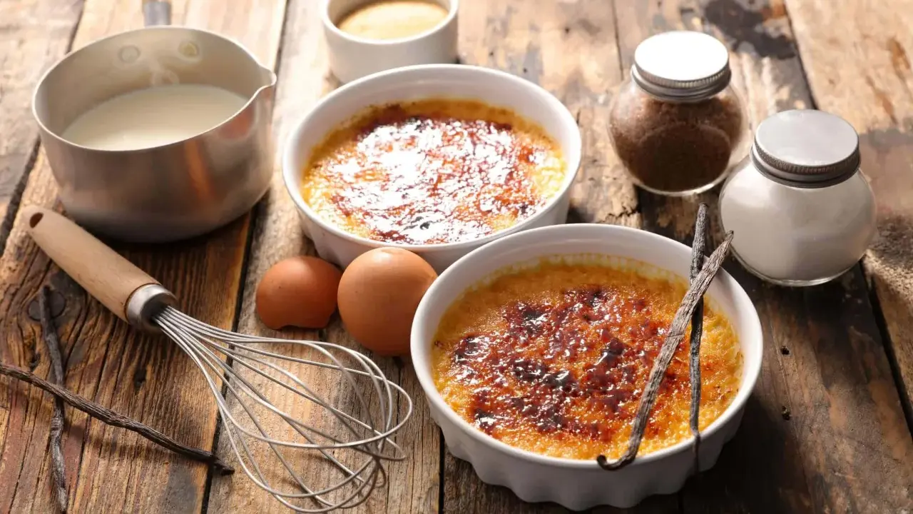 Dwa kremowe desery creme brulee z karmelizowanym cukrem, obok składniki: śmietanka, jajka, laski wanilii, cukier.