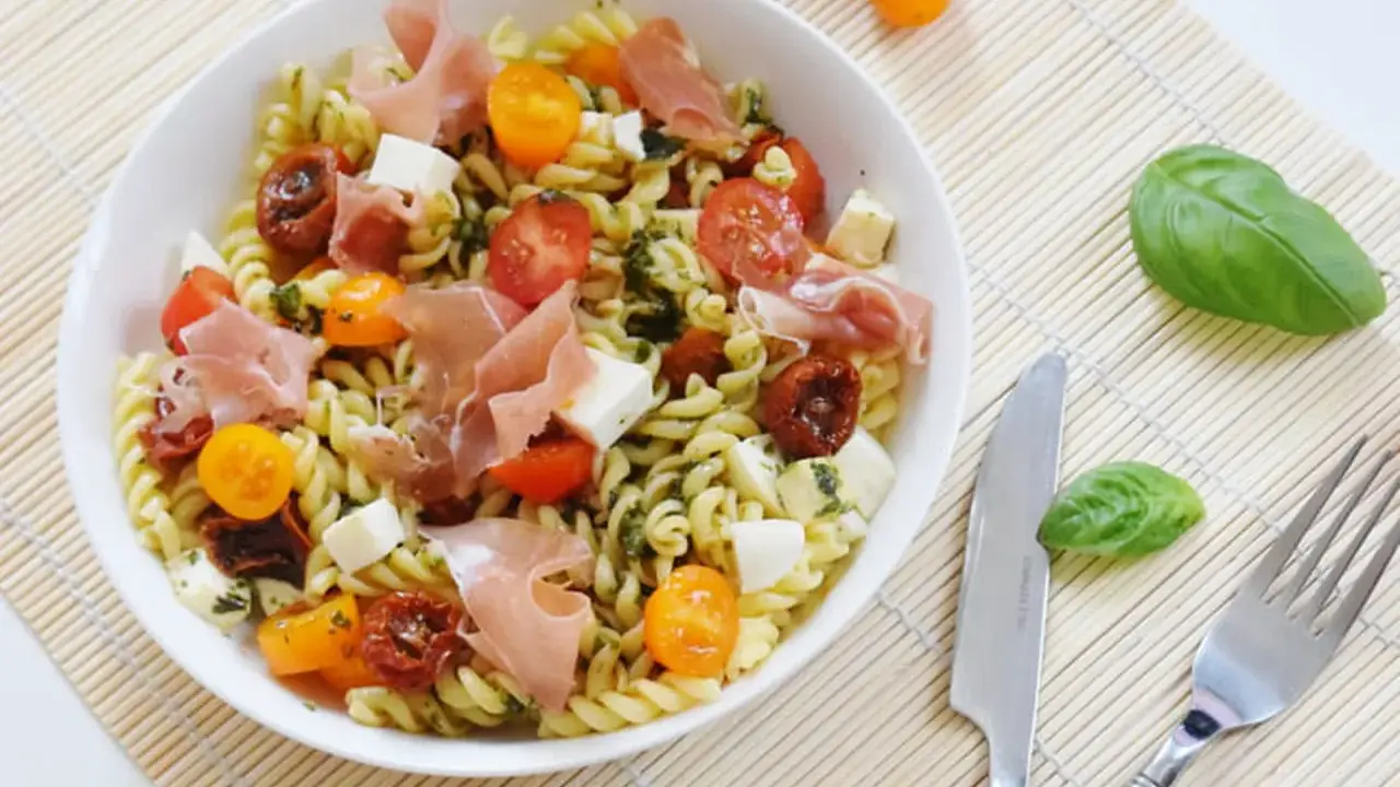 Une salade de pâtes originale avec des tomates cerises, du prosciutto, des dés de mozzarella et une sauce verte savoureuse.