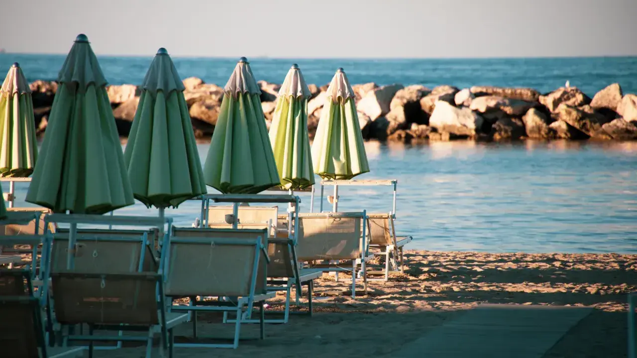 Ombrelloni verdi e lettini vuoti sulla spiaggia di una città riviera romagnola, con il mare calmo e una diga di sassi sullo sfondo.