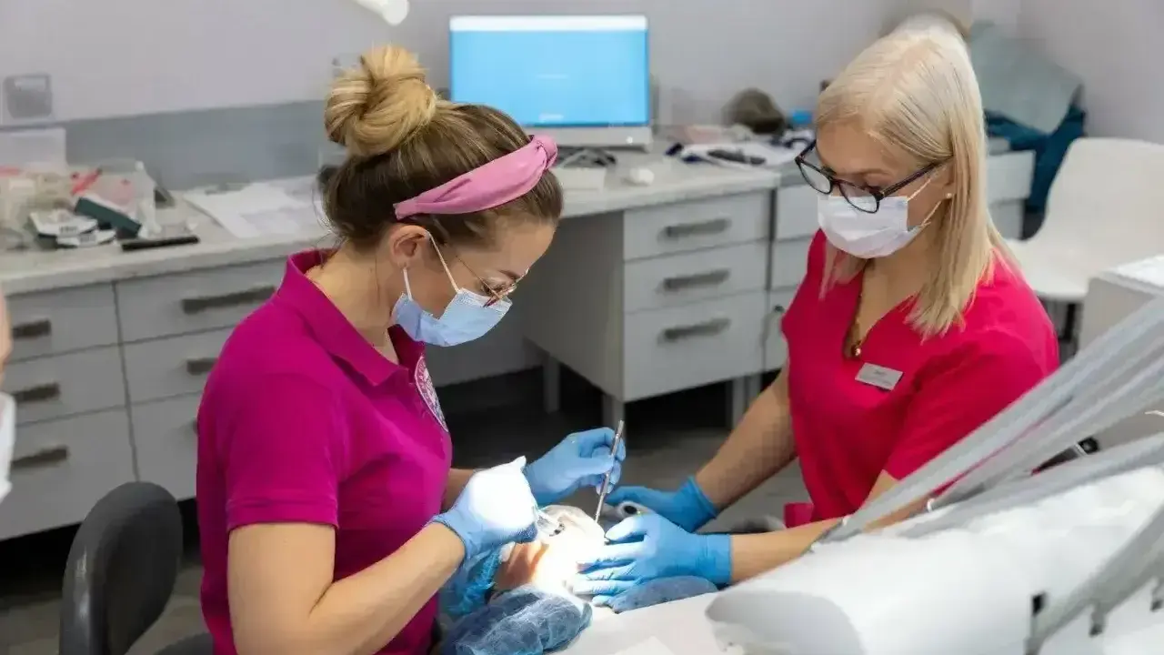 Dwie dentystki w maseczkach i rękawiczkach pracują przy pacjencie. Jedna z nich używa narzędzi, druga asystuje. Zabieg bez bólu dzięki lidokainie.